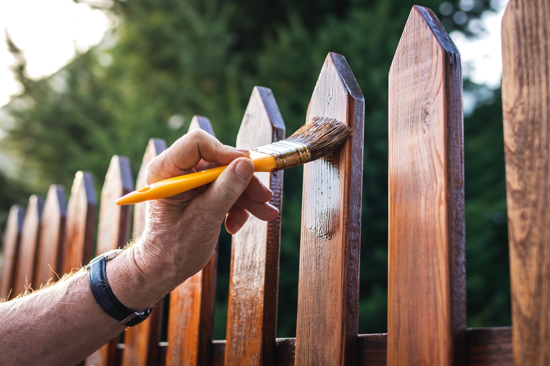 A painter using a paintbrush to stain a wooden picket fence in the backyard.