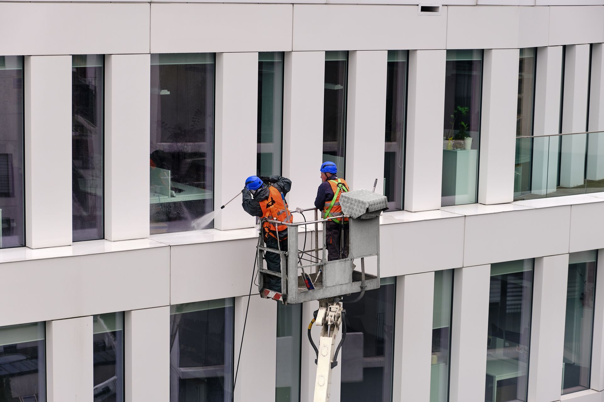 Two workers in safety harnesses power wash the exterior of a business building. They are standing in a crane cradle or aerial platform, utilizing a pressure washer and mops to clean the facade.