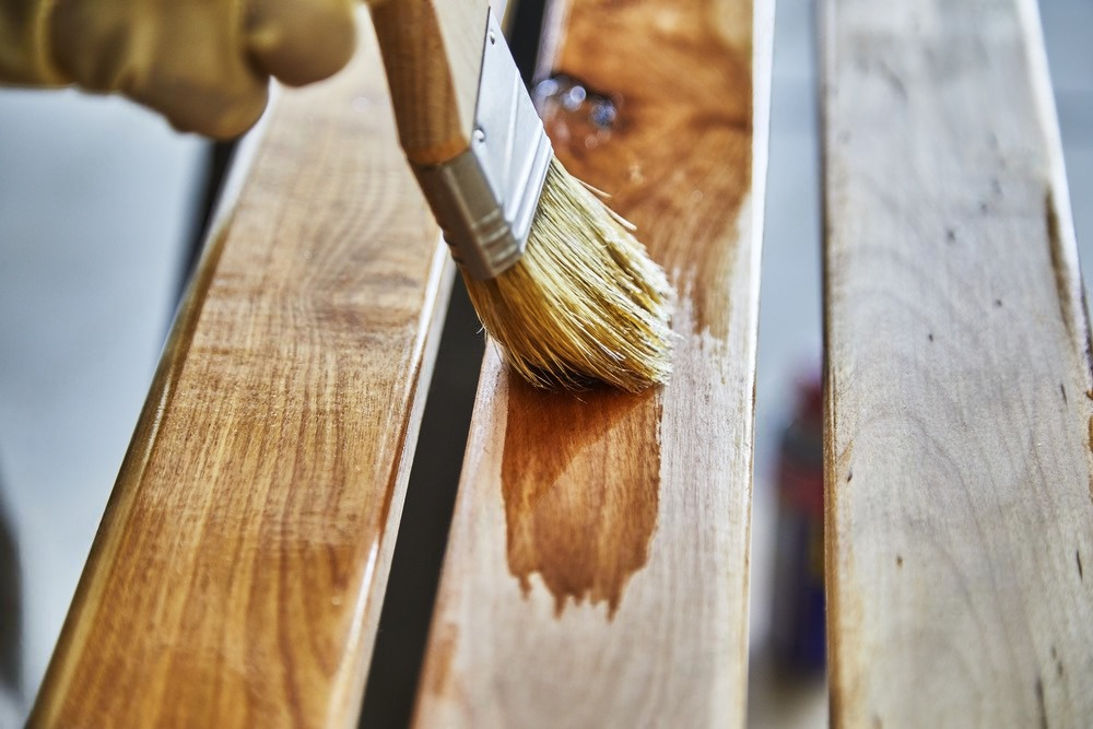 A person wearing gloves is applying wood stain to a piece of wood using a brush.