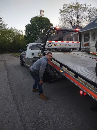 Man in jeans and gray hoodie looks at a flatbed tow truck, in front of a house, on a driveway.