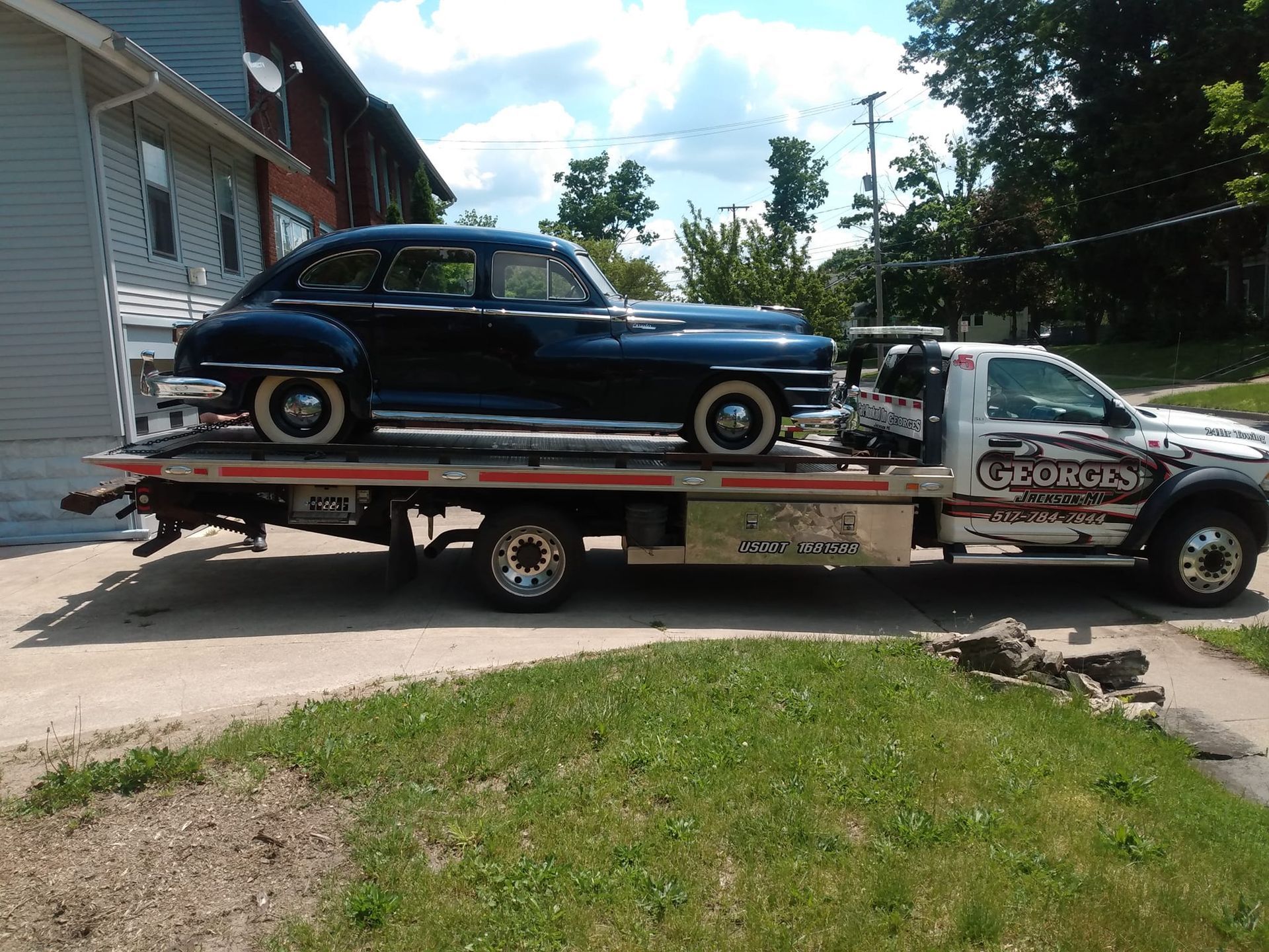 Black vintage car on a tow truck in front of a house, under a cloudy sky.