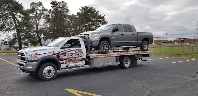 Tow truck carrying a gray pickup truck in a parking lot on a cloudy day.