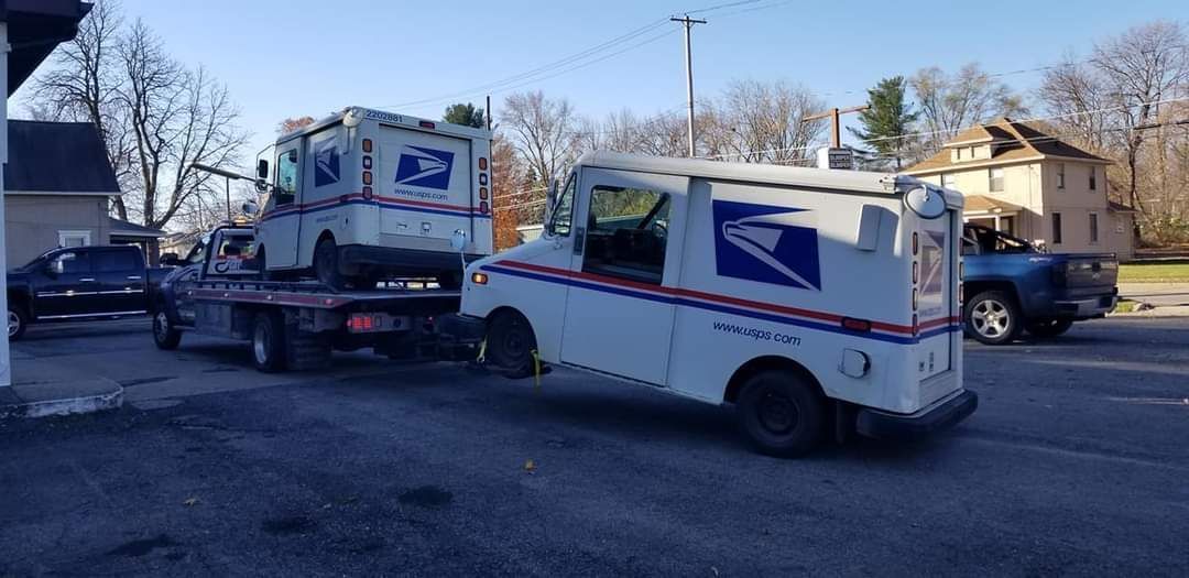 Two USPS mail trucks; one driving and one on a flatbed tow truck in a parking lot.