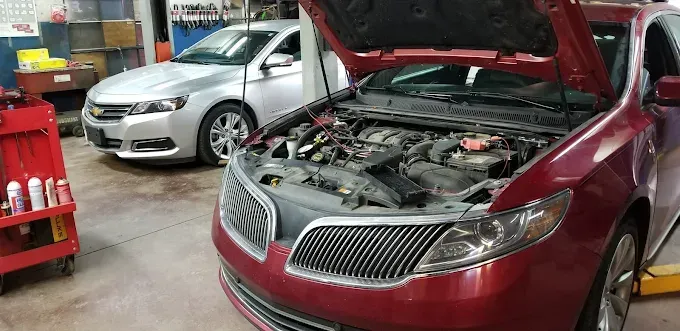 A red car with an open hood sits in a garage next to a silver car, with tools visible.