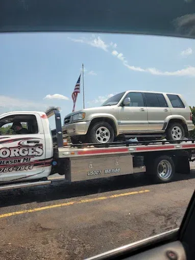 A beige SUV being towed on a flatbed truck on a sunny day. An American flag flies in the background.
