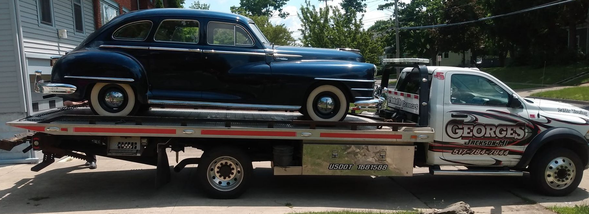 A classic dark blue car is being transported on a tow truck in an urban setting.