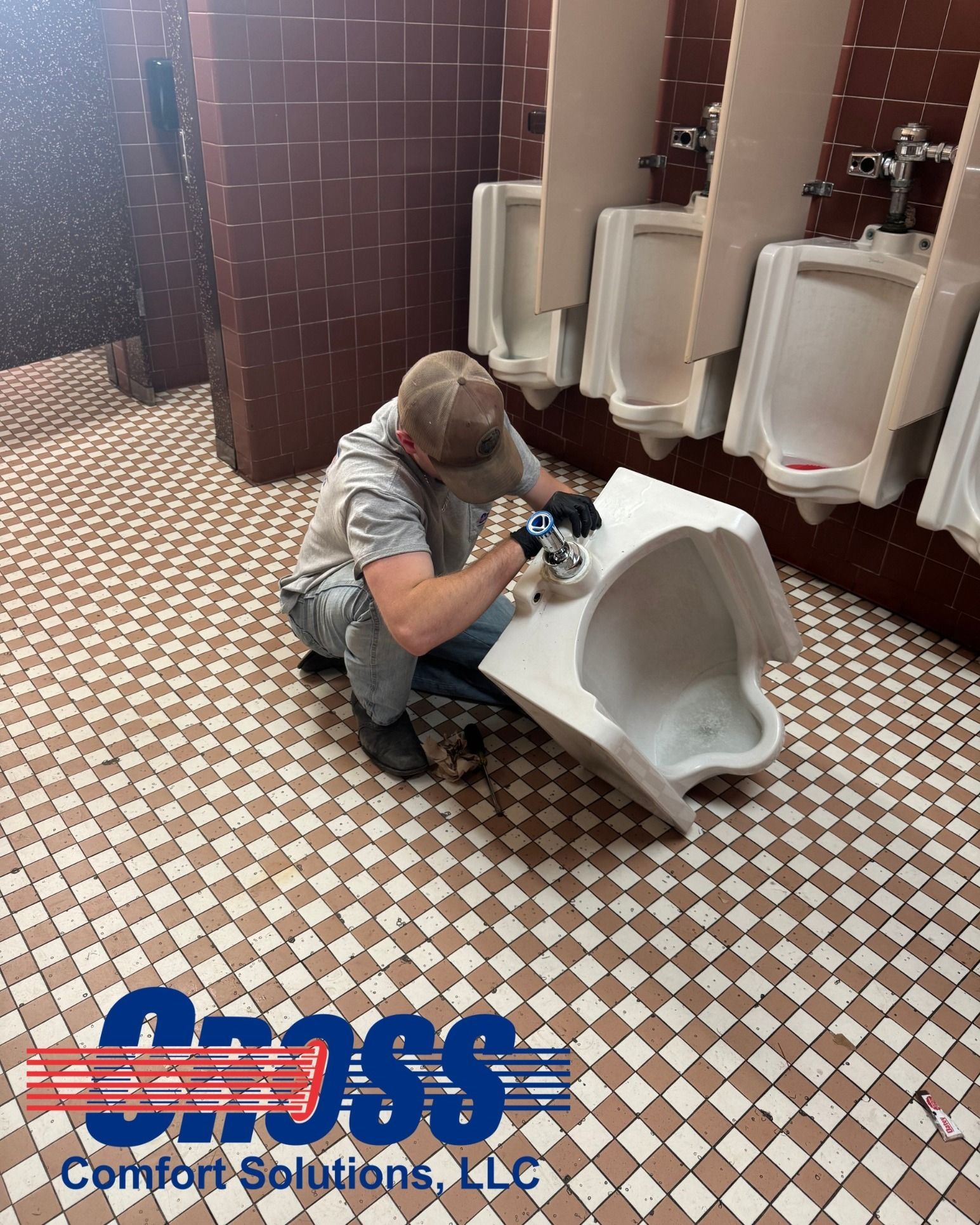 Man installing a urinal in a public restroom with red tile walls and checkered floor.