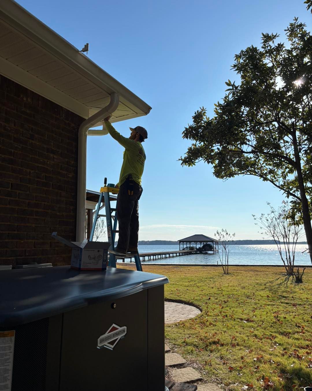 Person on ladder installing gutter on a brick house by a lake on a sunny day.