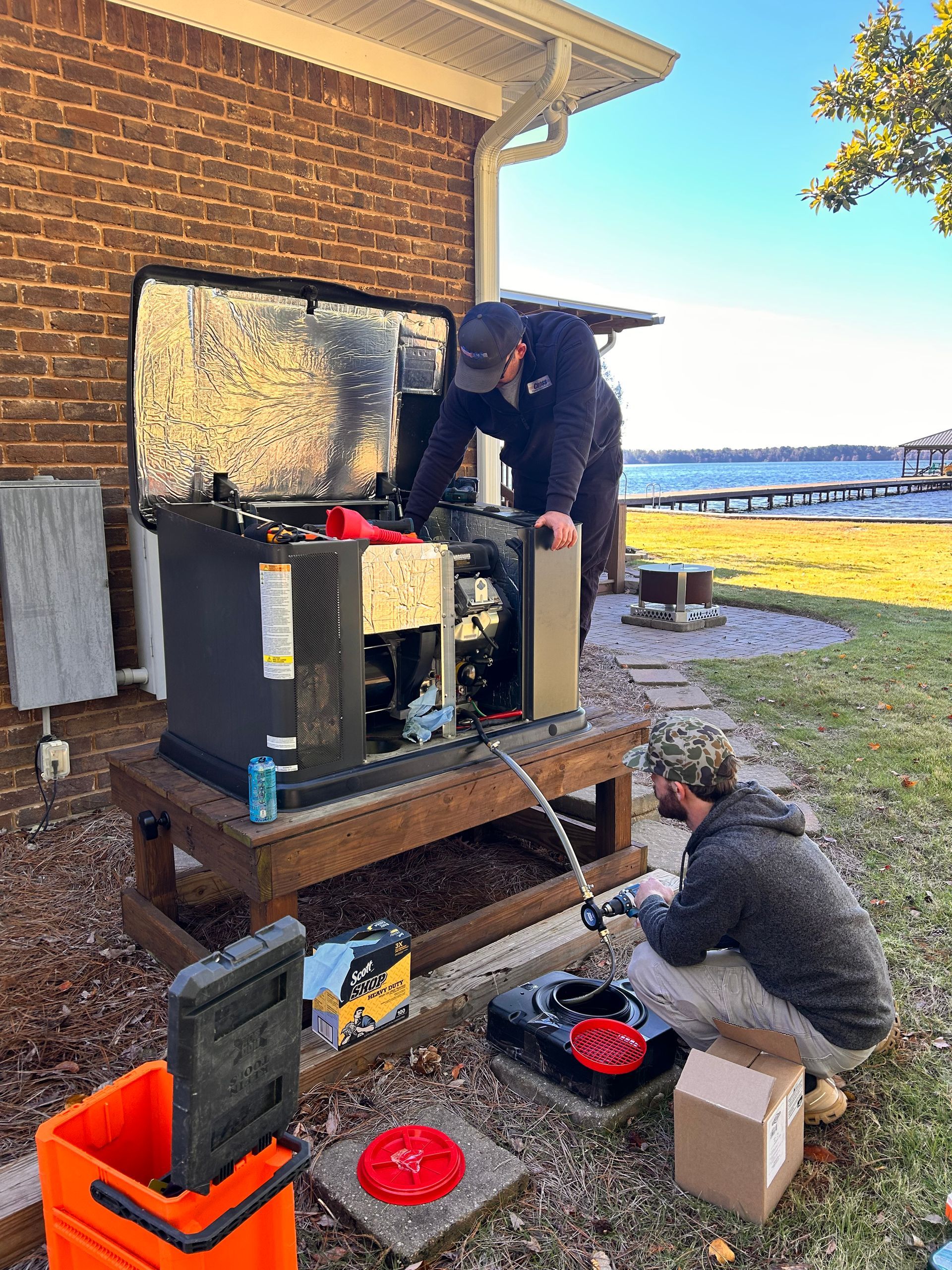 Two people working on a generator outside a brick building near a lake. One kneels, the other leans in.