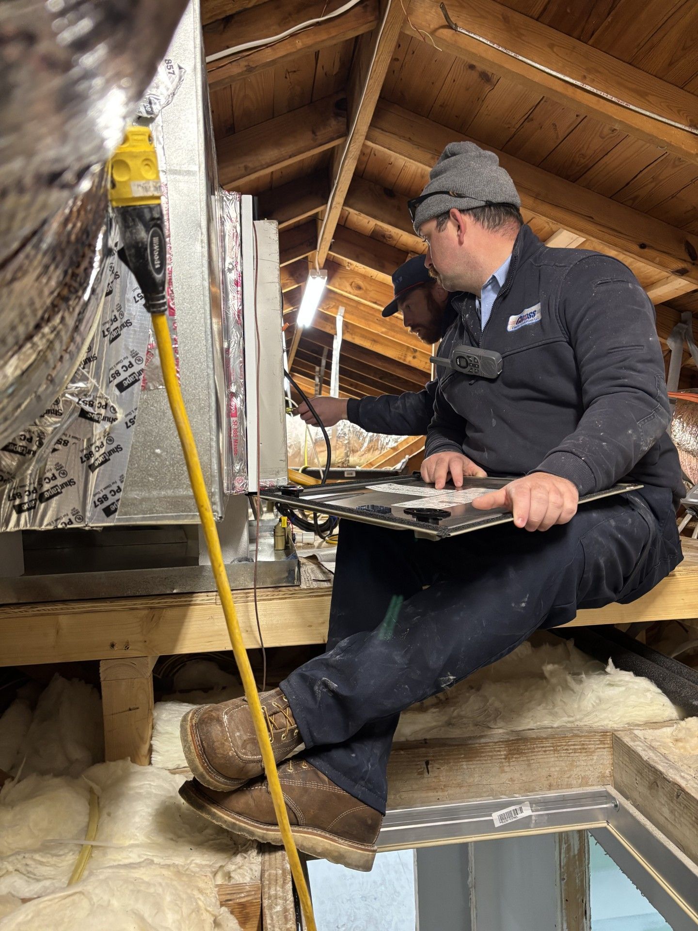 Two technicians inspecting an HVAC unit in an attic. One holds a clipboard; yellow extension cord in view.