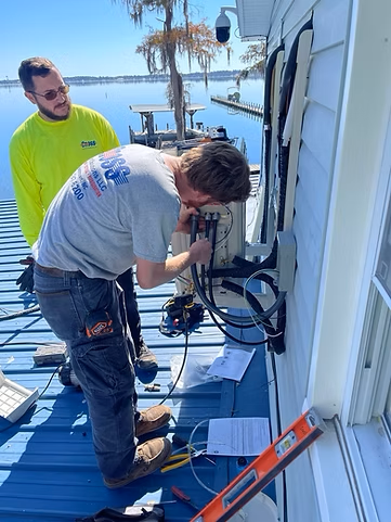 Two men are working on an air conditioner on the side of a house.