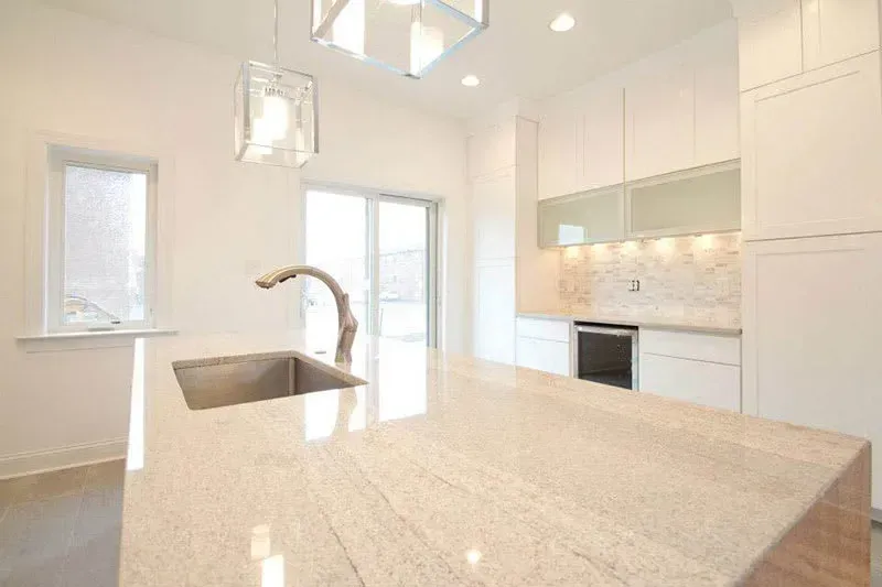 Modern white kitchen with a granite countertop island, sink, and cabinets. Sliding door and window provide natural light.