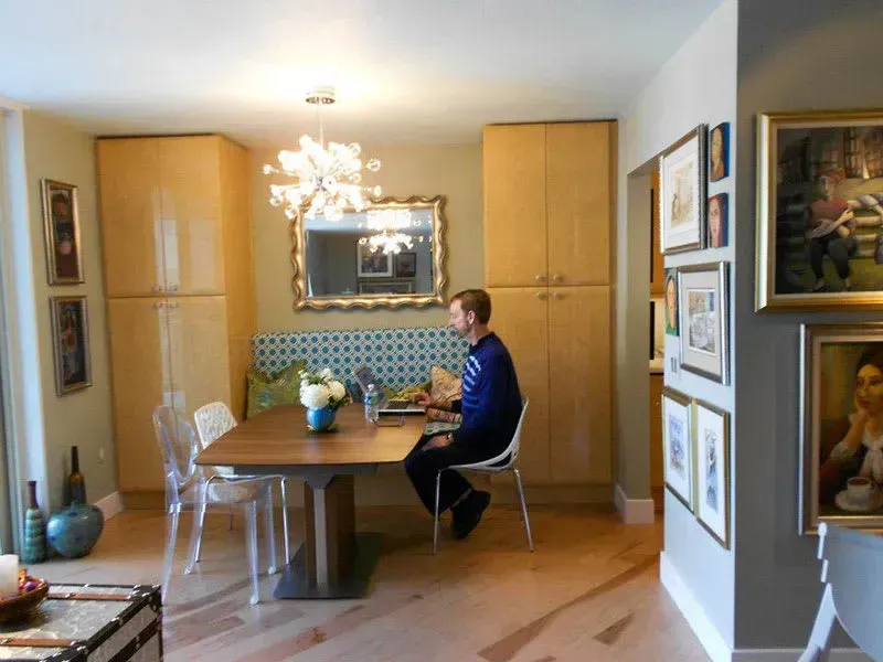 Man sits at dining table, working on a laptop. Light wood cabinets, framed art, and a chandelier are visible.