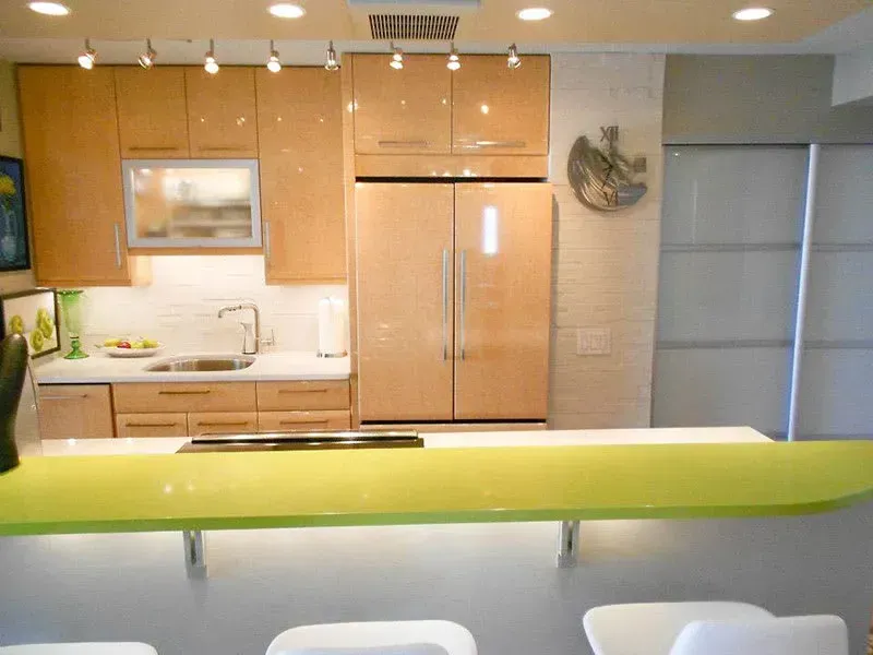 Modern kitchen with light wood cabinets, green countertop, and stainless steel refrigerator.