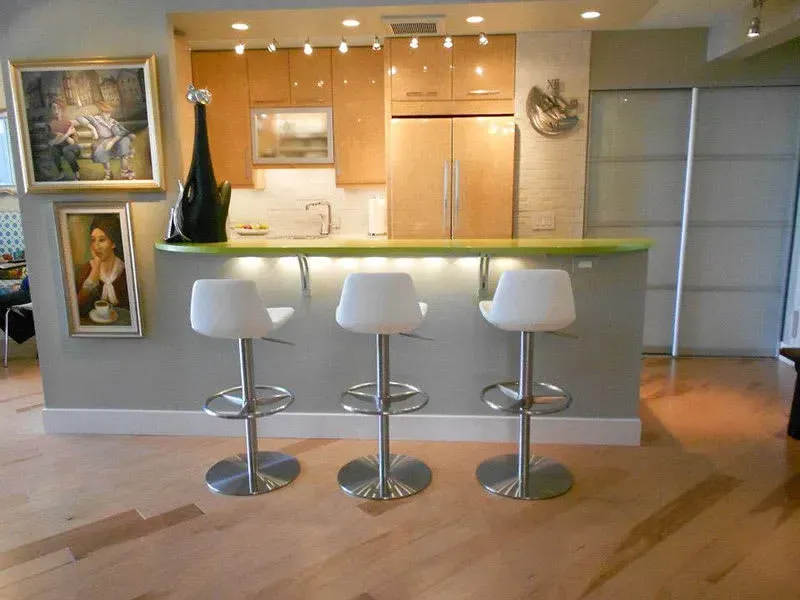 A modern kitchen with a bar counter and three white stools. Wooden cabinets, artworks, and a sliding door.