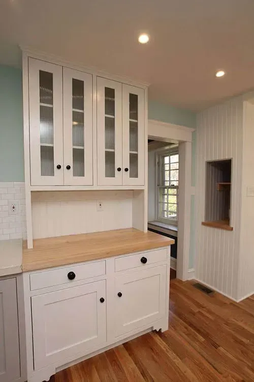 White kitchen cabinet with glass doors, wood countertop, and black knobs; light blue wall.