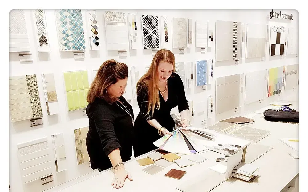 Two women reviewing paint swatches in a design showroom. Displayed samples on wall.