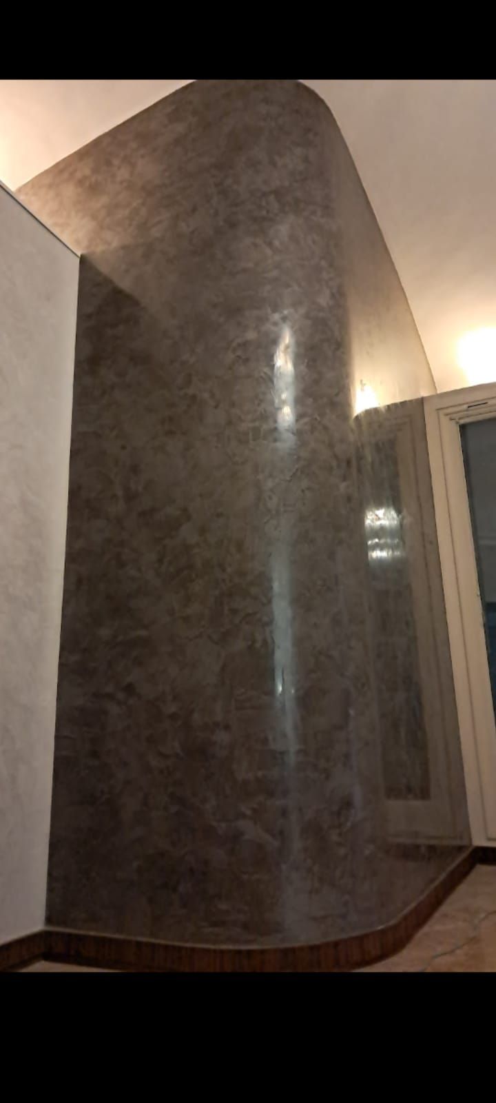 Una parete curva e ruvida con una finitura maculata scura incontra un soffitto bianco. È visibile una porta con cornice in legno.