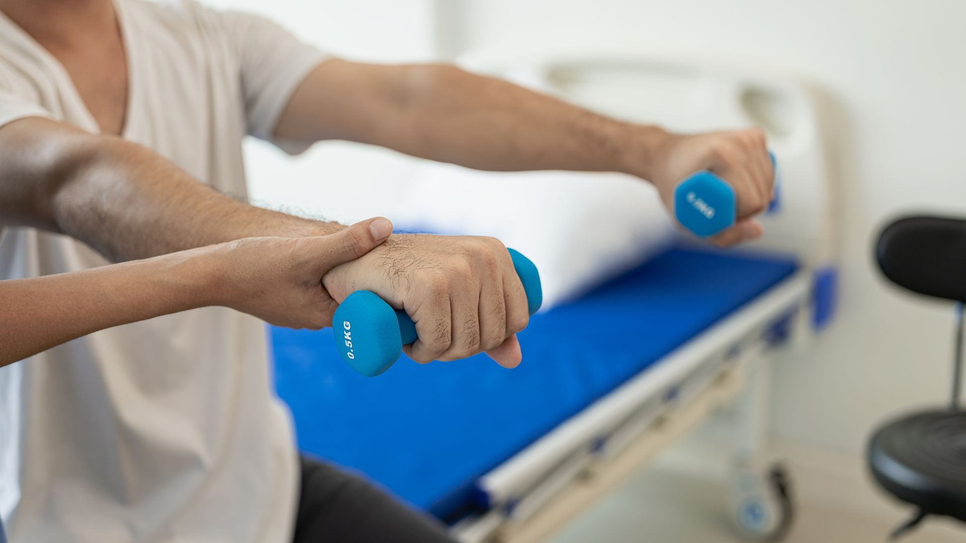 Person exercises with dumbbells, assisted by another person, near a hospital bed.