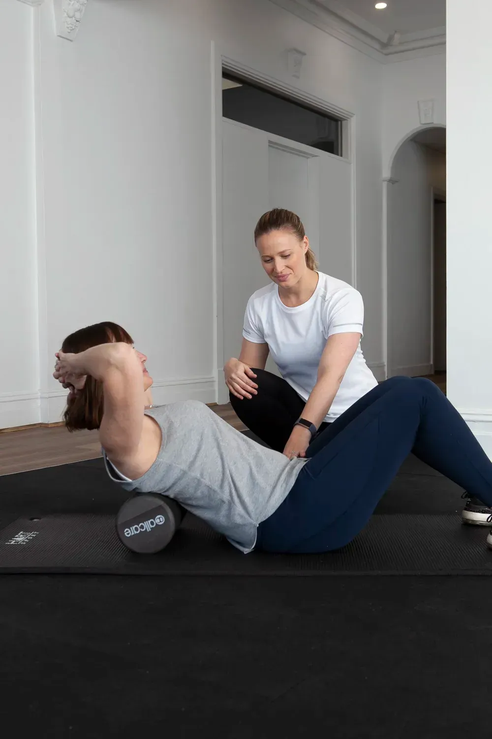 Woman using foam roller for back, supervised by another woman in a bright room.