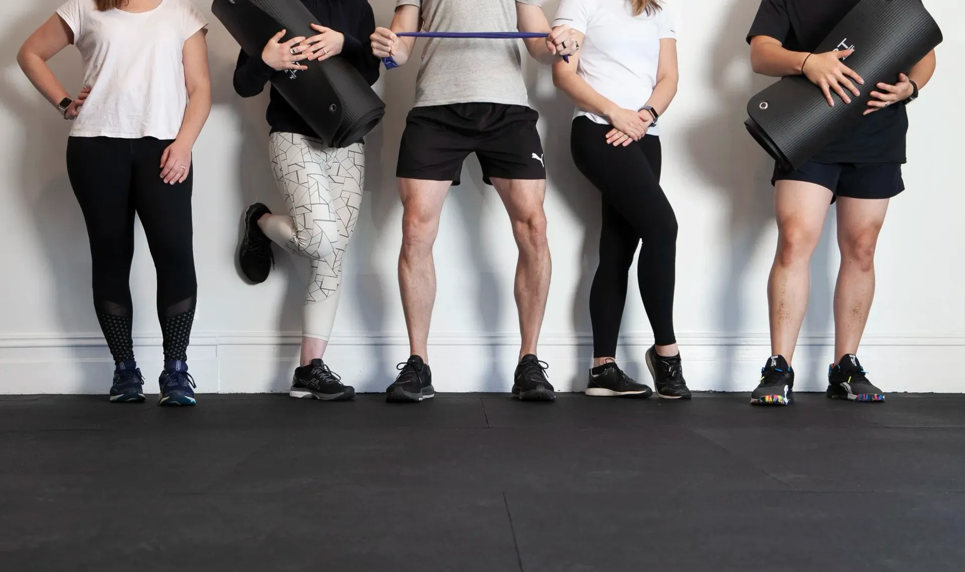 Five people in workout clothes, holding equipment, standing against a white wall and dark floor.