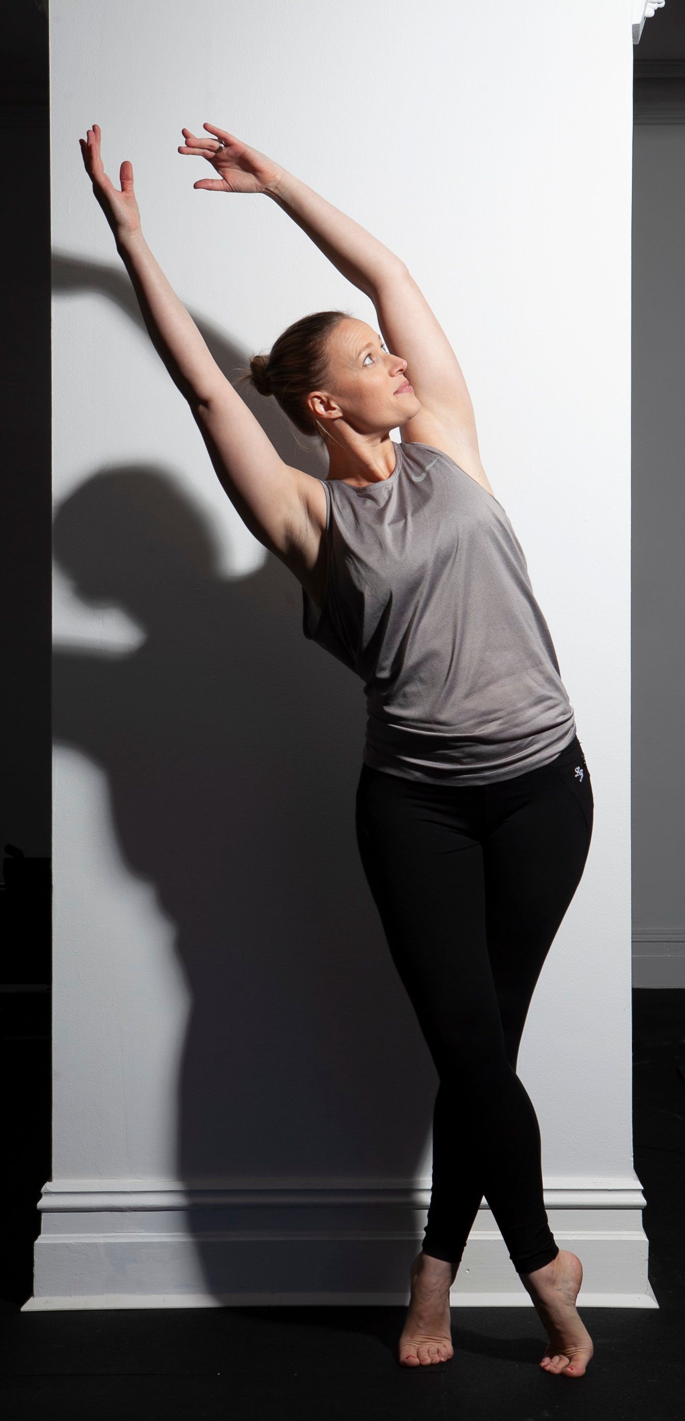 Woman in athletic clothing leaning against a white wall, arms raised. Shadow cast on the wall.