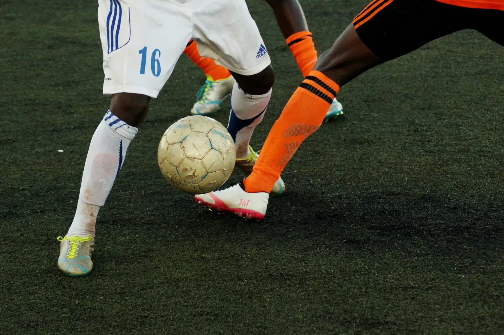 Two soccer players in action on a green field, kicking the ball. One in white, the other in orange.