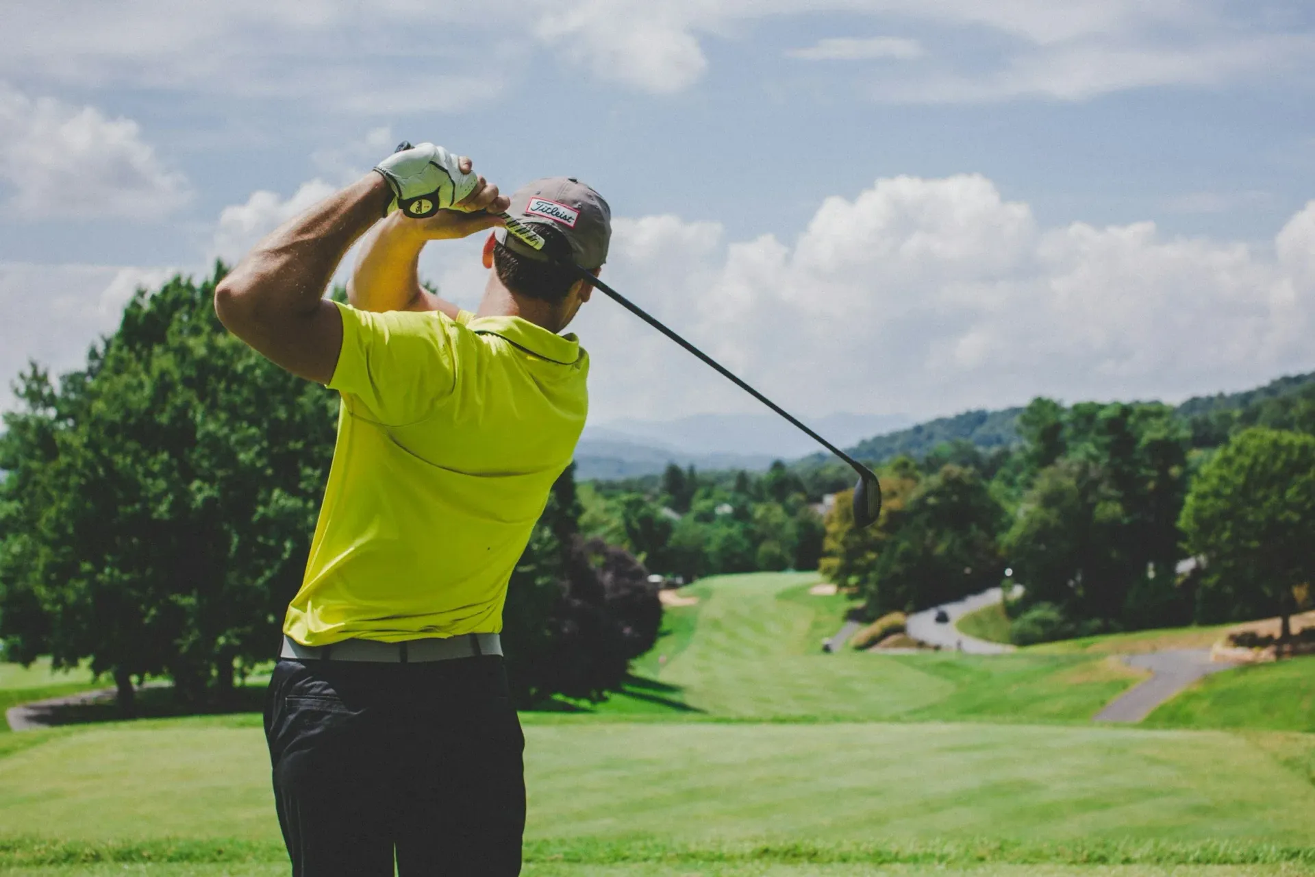 Golfer in a yellow shirt swinging a club on a green golf course with trees and mountains in the background.