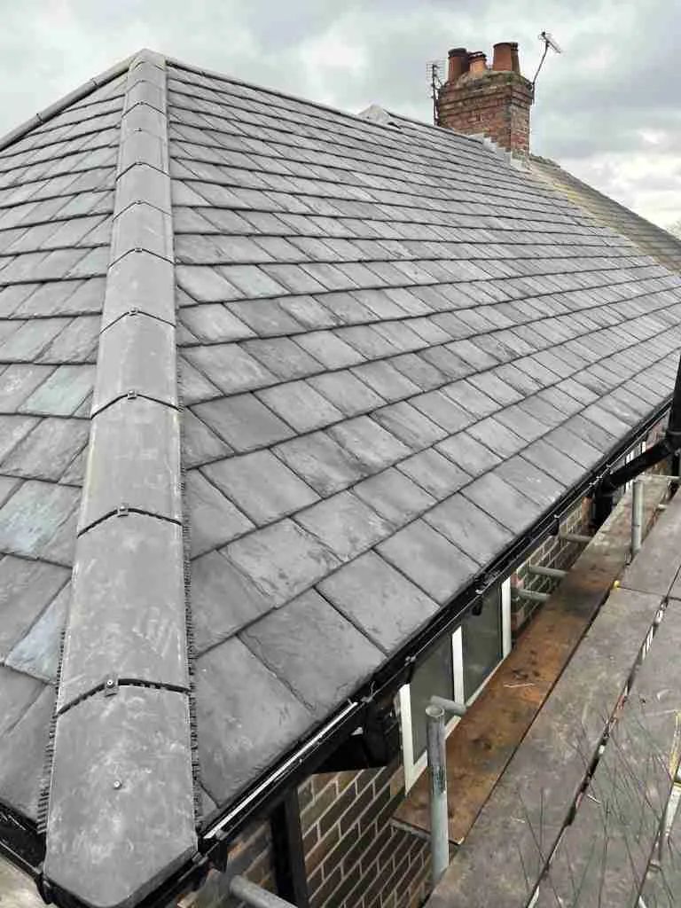 Dark gray slate roof of a house with a brick chimney and guttering visible. Cloudy sky.