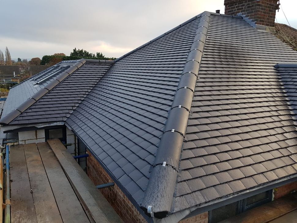 Dark gray tiled roof on a house with a brick chimney and skylights under a cloudy sky.
