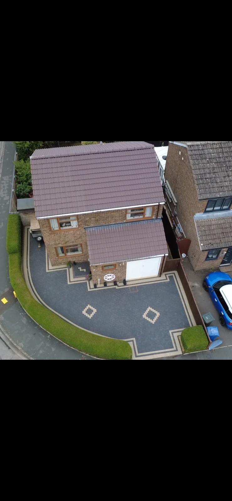 Overhead view of a brick house with a paved driveway and green hedges. A blue car is parked nearby.