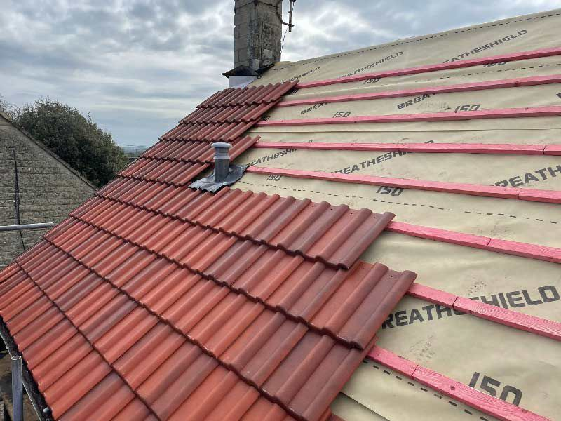 Red roof tiles being installed on a house roof with insulation and chimney visible.
