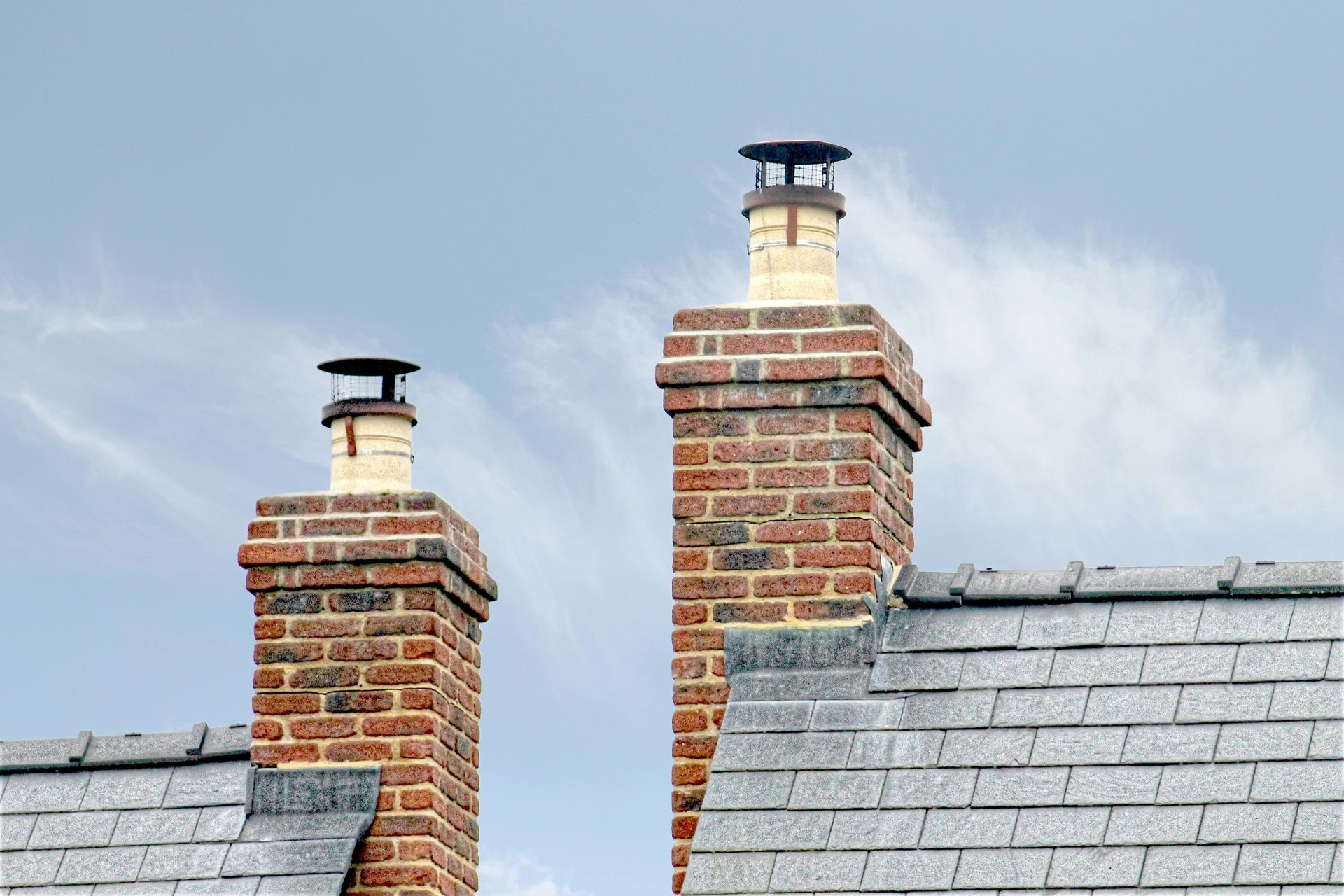 Two brick chimneys on a gray shingle roof against a cloudy blue sky.