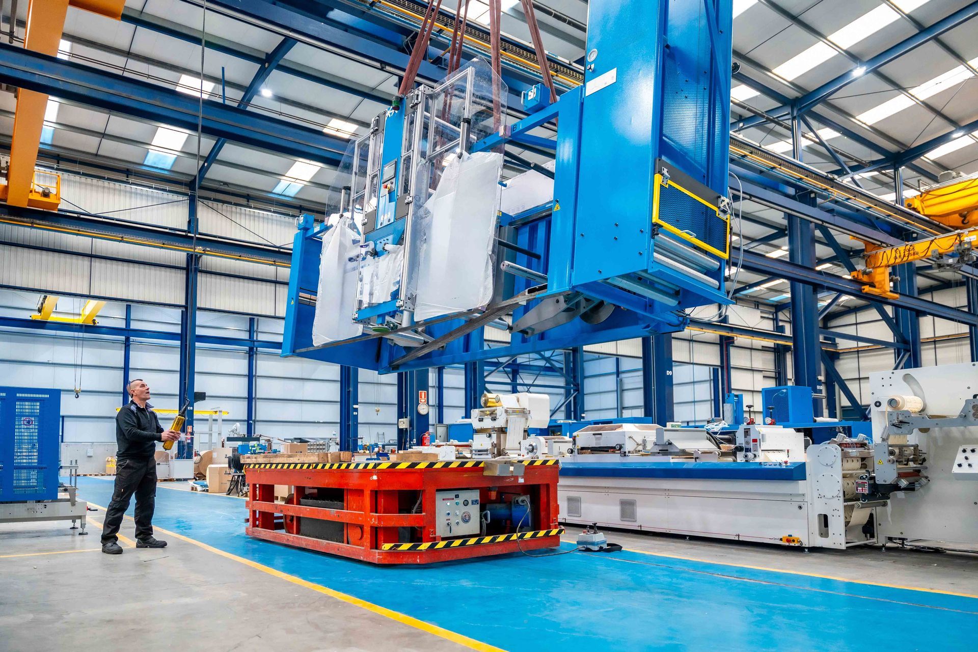 Worker guides a large blue industrial machine being lifted inside a spacious factory.