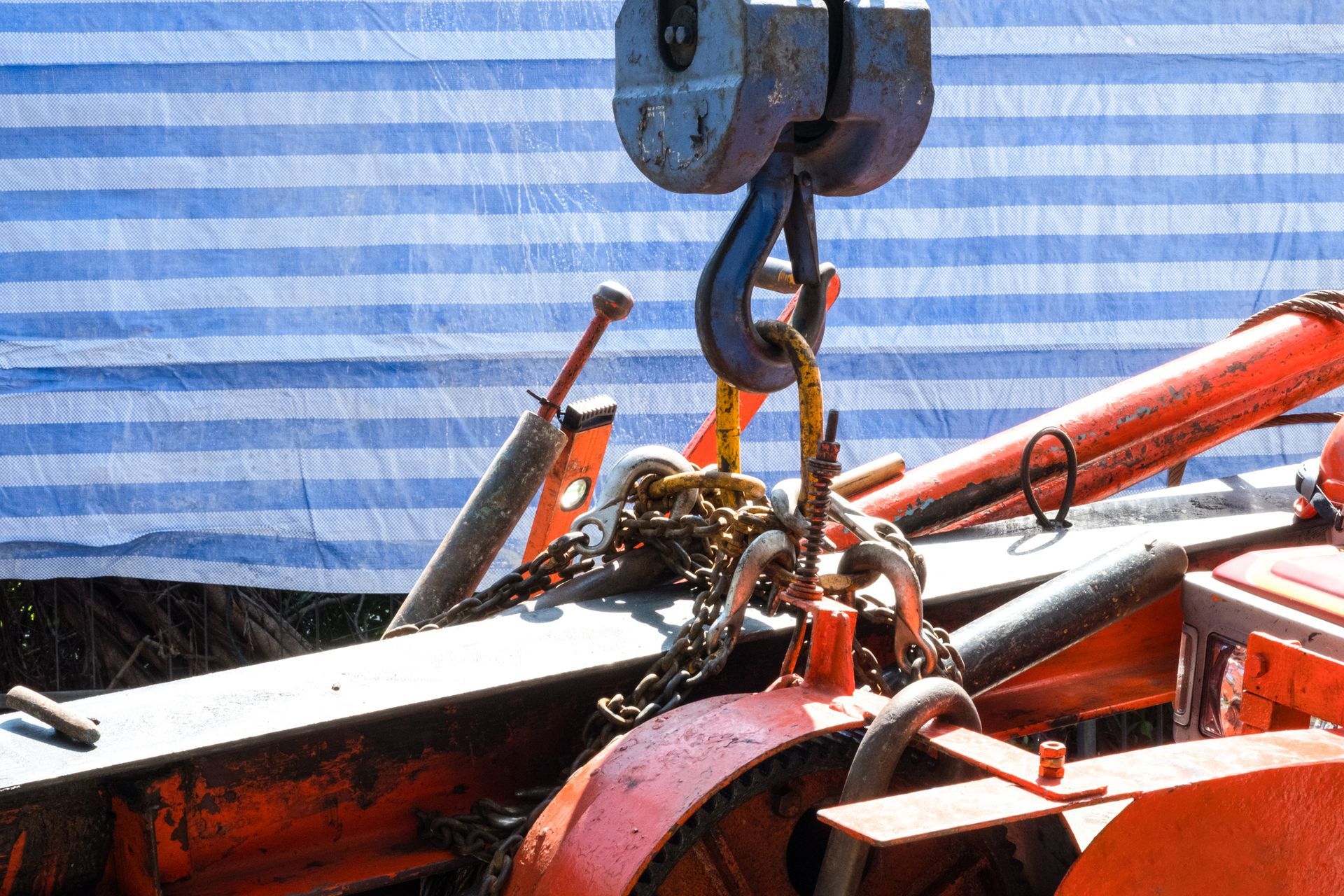 A crane hook lifting orange machinery with chains attached; blue and white striped tarp in background.