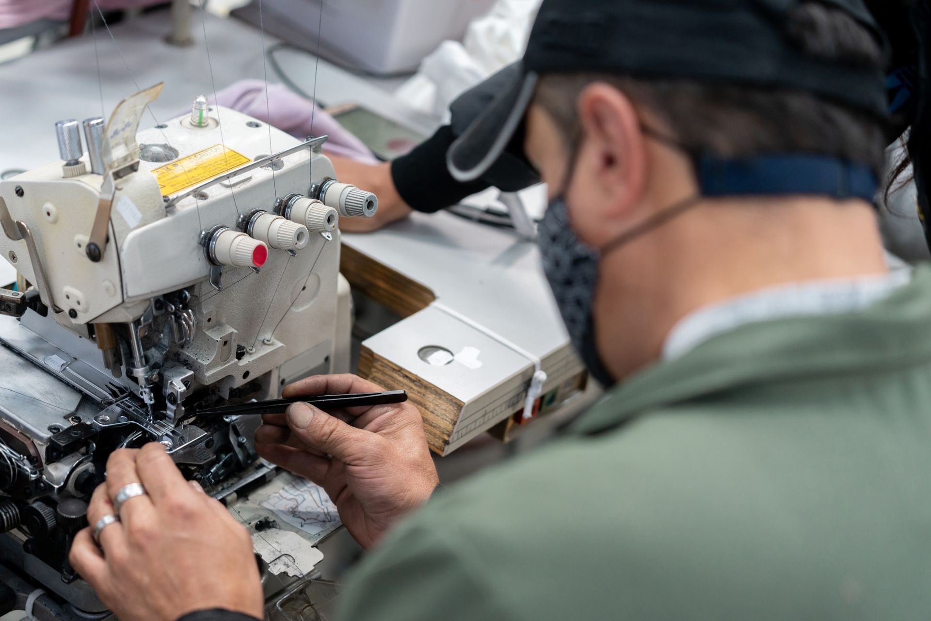 A person in a mask and cap repairs machinery, holding tweezers. Workspace with a sewing machine.