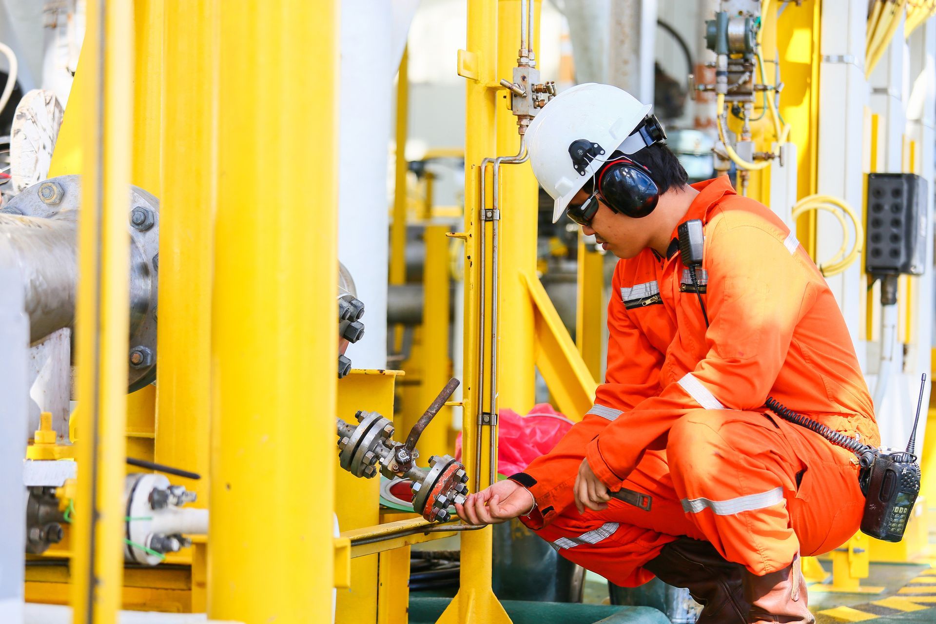 Oil worker in orange jumpsuit and hard hat inspecting equipment on an offshore platform.