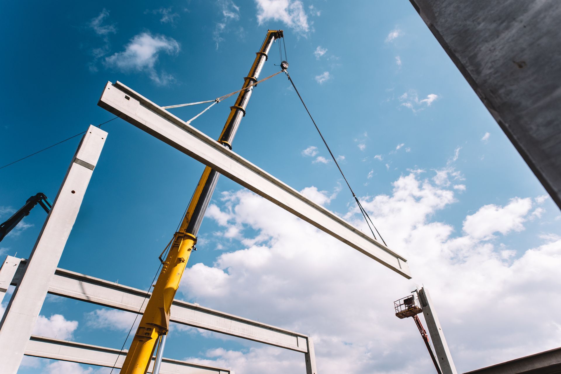 Crane lifting a concrete beam at a construction site against a blue sky with clouds.