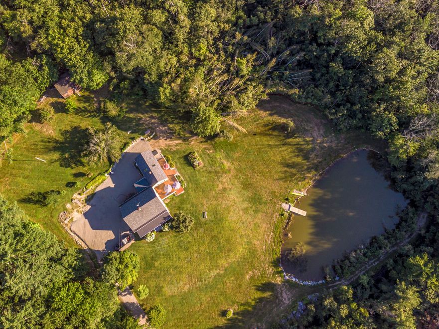 Aerial view: house with a long driveway, large grassy yard, pond with dock, and surrounding trees.