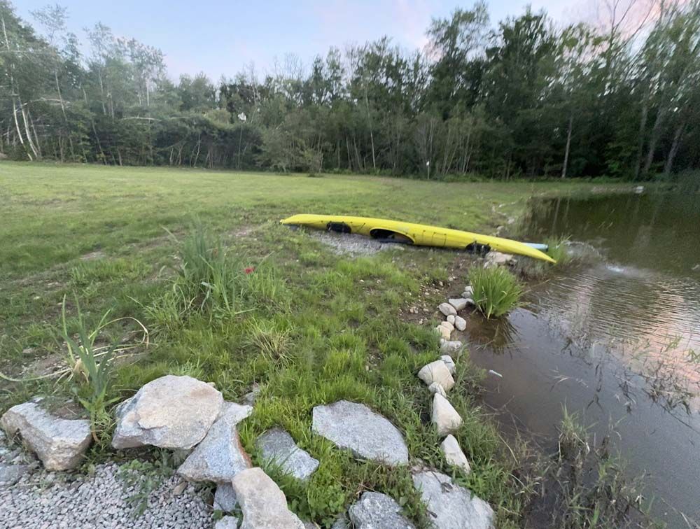 Yellow kayak on grassy bank near pond with rocks and trees.