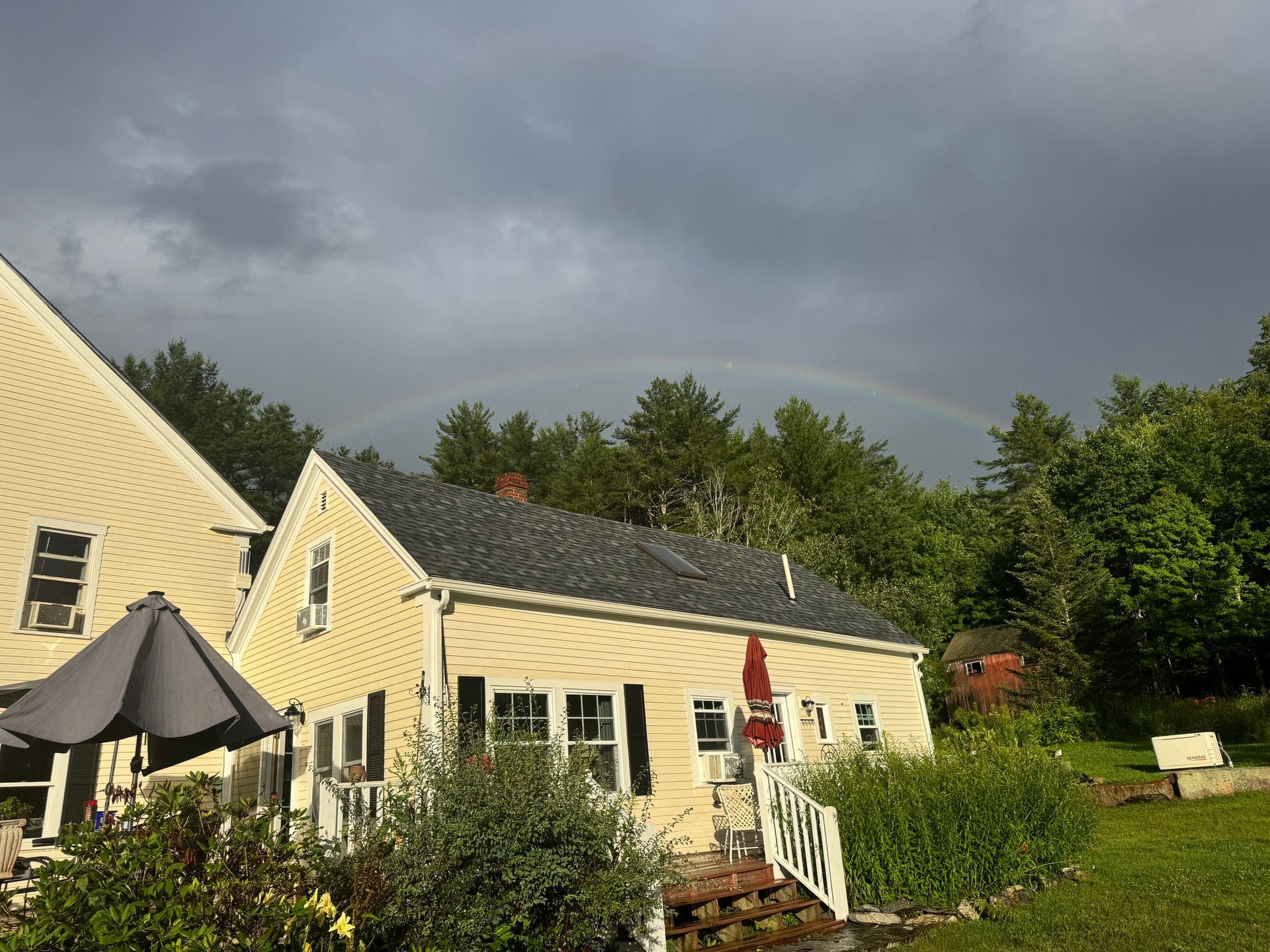 Yellow house with a rainbow arching above the roof and treeline. Dark clouds and greenery surround the scene.