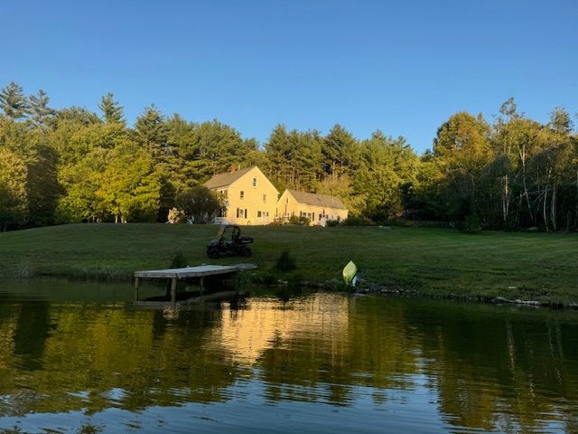 House reflected in a pond; green lawn, trees, blue sky, and a small dock.