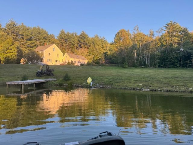 A house and pond are reflected in the water. A dock and tractor sit on the grass. Trees frame the scene.