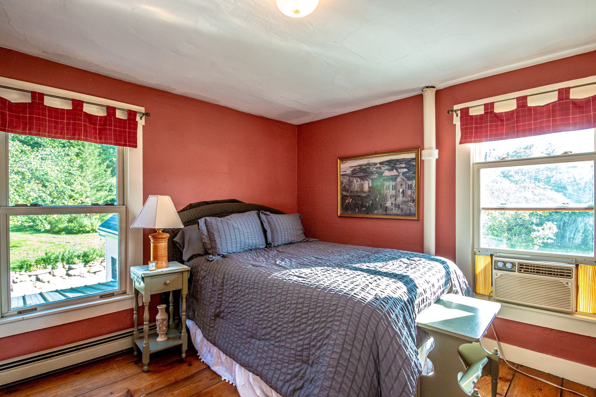 Bedroom with red walls, bed with blue quilt, two windows with red shades, and a framed picture.