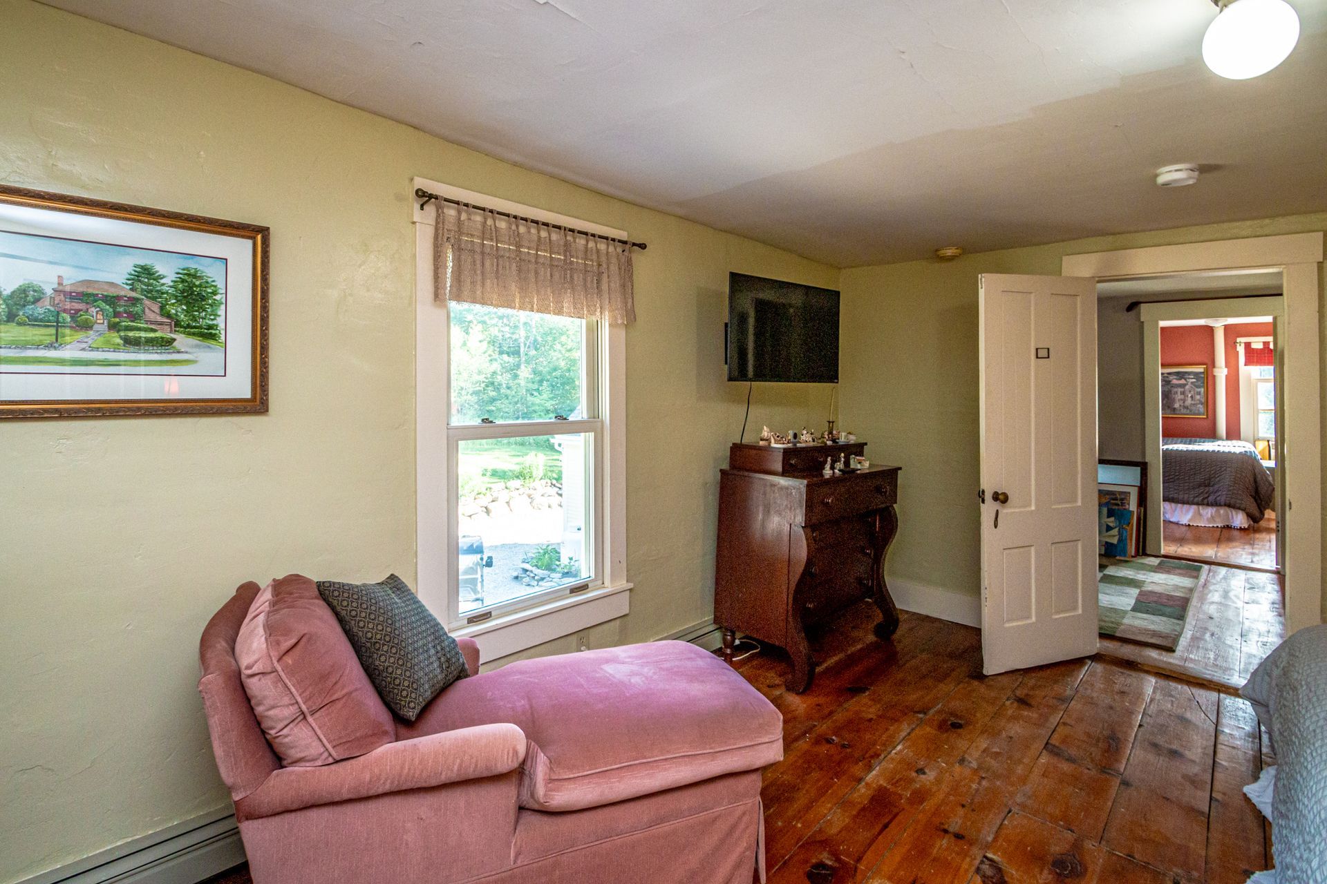 Bedroom with pink chaise lounge, wooden dresser, window, and doorway to another room.