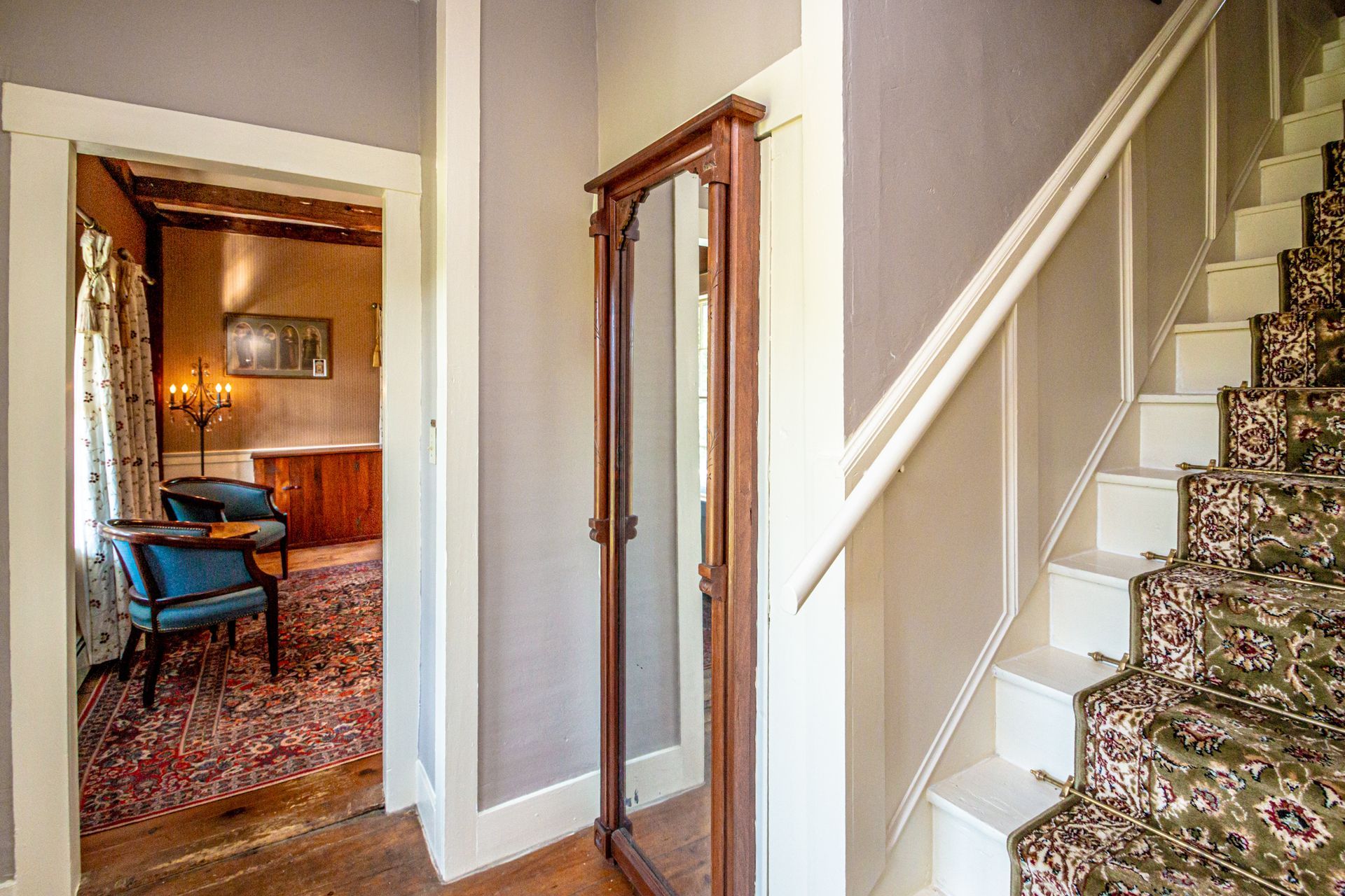 Entryway with staircase, full-length mirror, doorway to room with furniture, and patterned carpet.