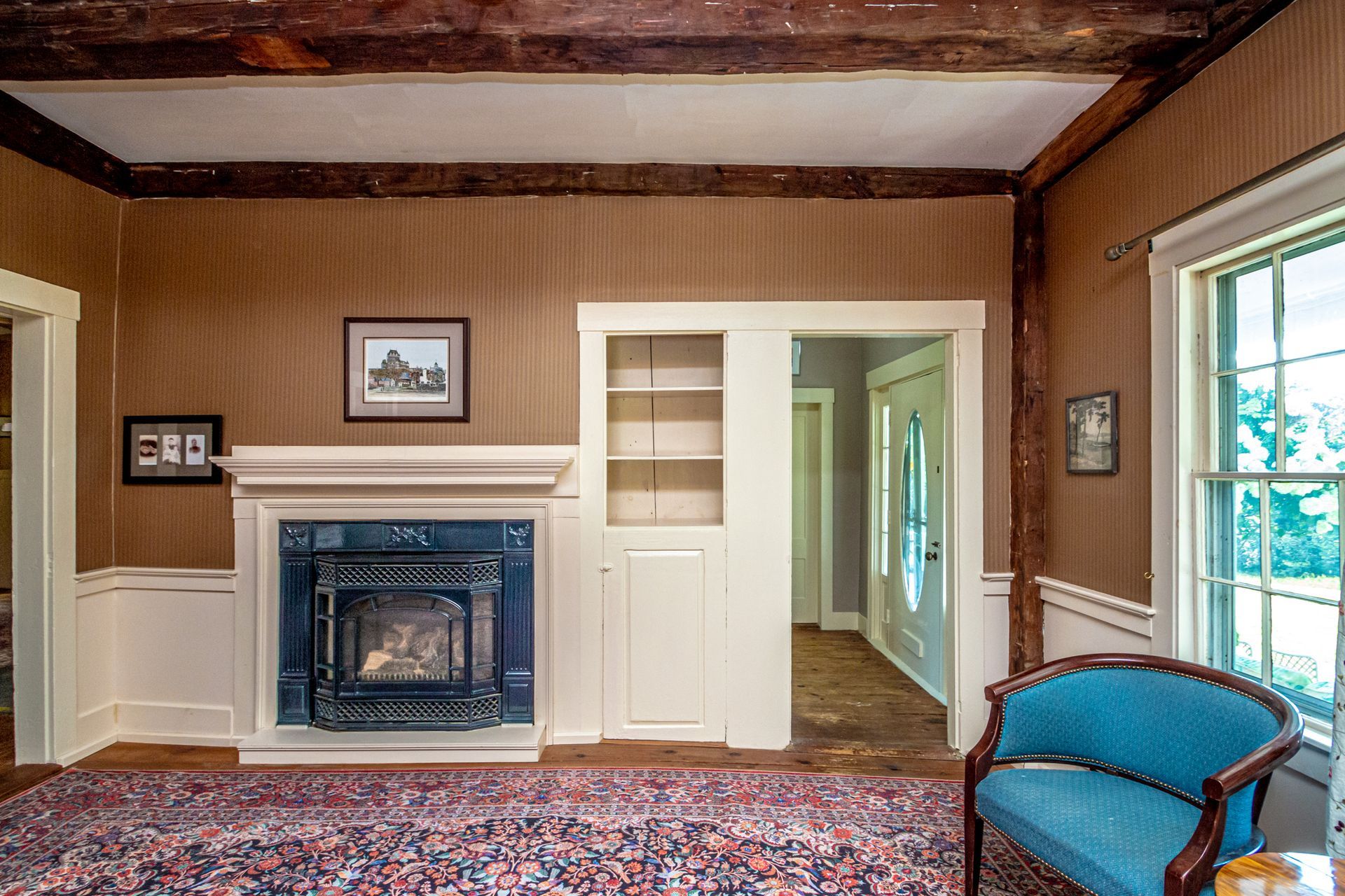 Living room with fireplace, built-in shelving, blue chair, and rug. Brown walls with exposed beams and white trim.