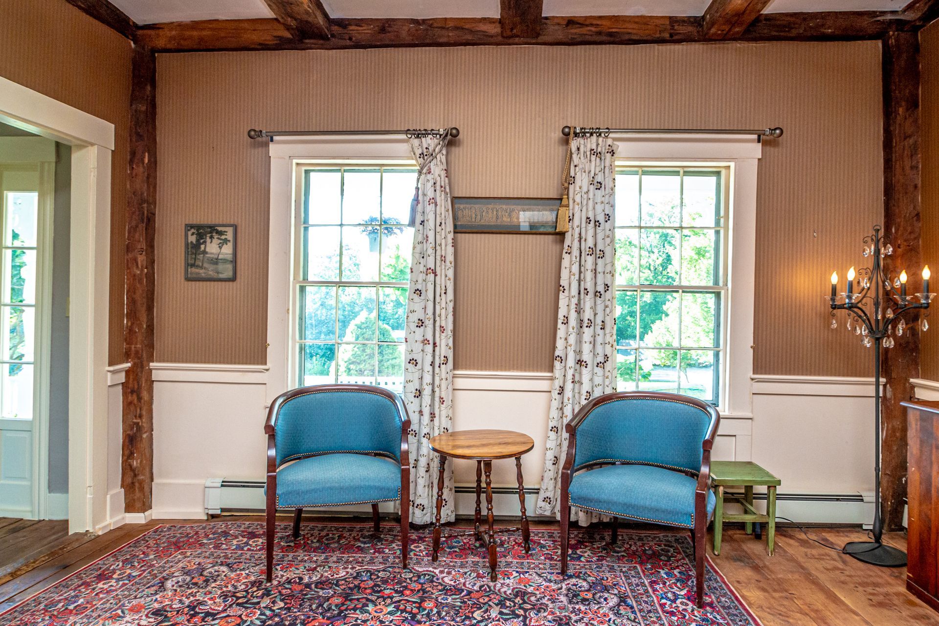 Room with two blue chairs, windows, a small table, and a rug. Beige walls, wooden beams, and a candle holder.