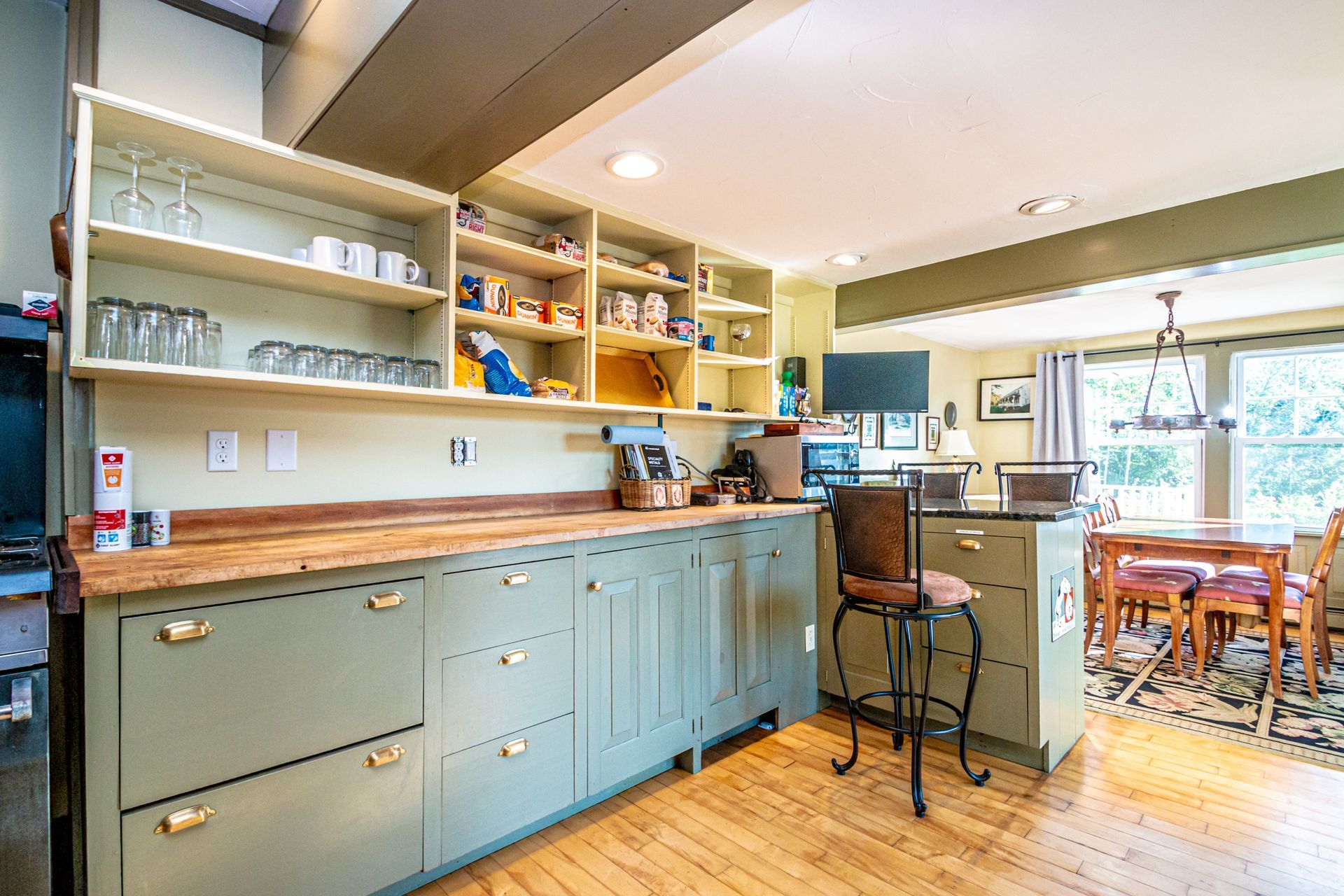 Kitchen with sage green cabinets, wooden countertops, and open shelving. Dining area visible.