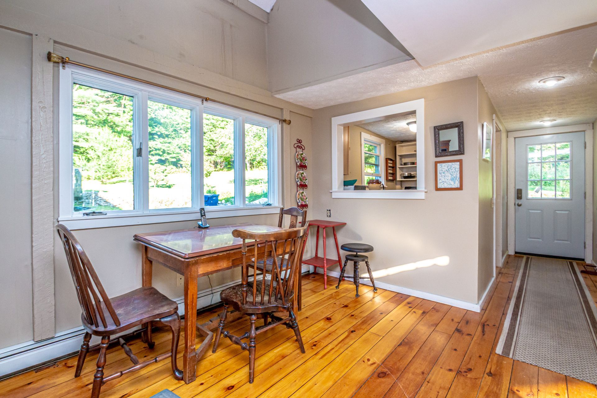 Dining room with wooden table, chairs, large window, and doorway to a hallway, with natural wood floors.
